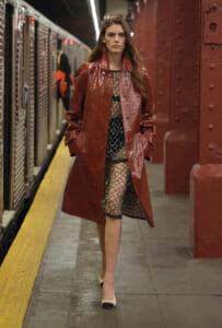 Woman in a red crocodile-embossed trench coat posing on a subway platform, wearing a sheer black fishnet dress with green accents and beige/black heels.