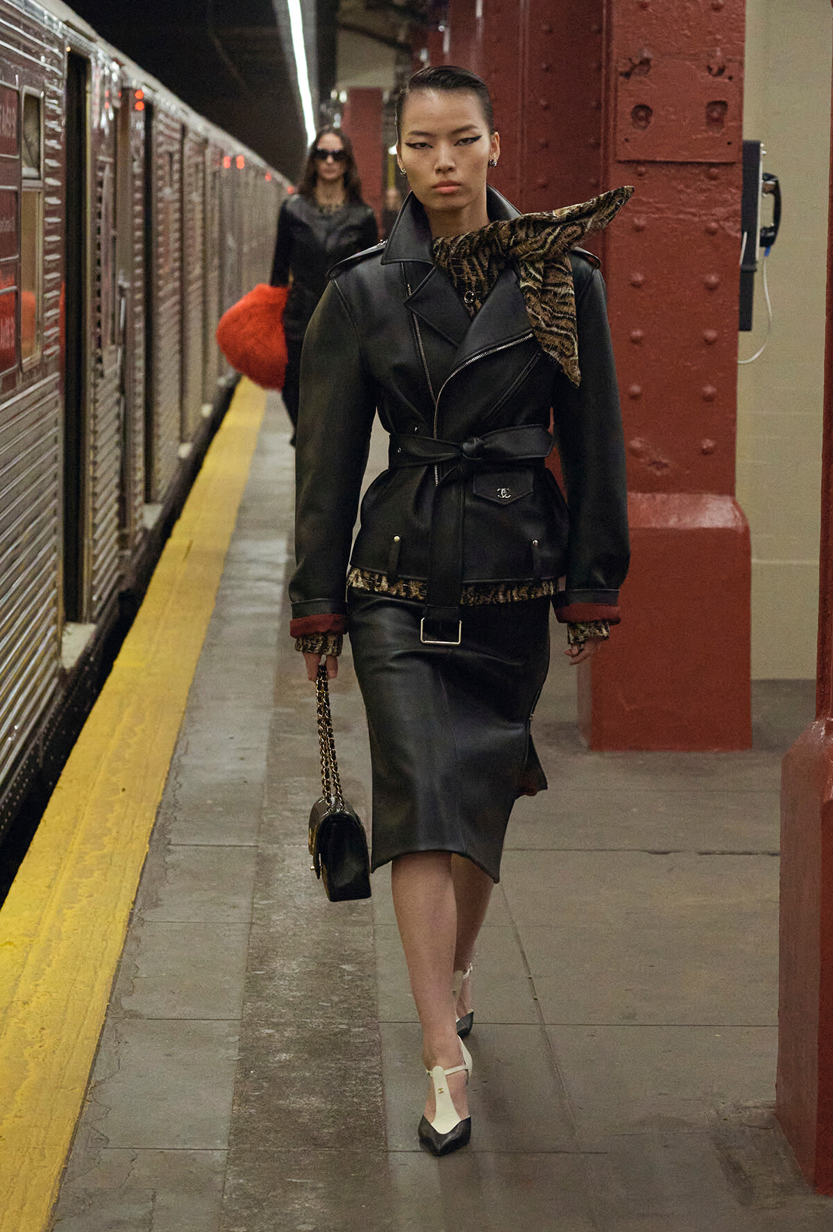 Woman in a black leather belted coat with a leopard-print scarf walking on a subway platform beside a train, carrying a small black purse with a chain strap.