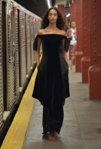Model in an off-the-shoulder black dress walking along a subway platform beside a train.