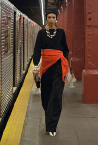 Woman wearing a black outfit with an orange sash walking on a subway platform, headwrap and chunky pearl jewelry.