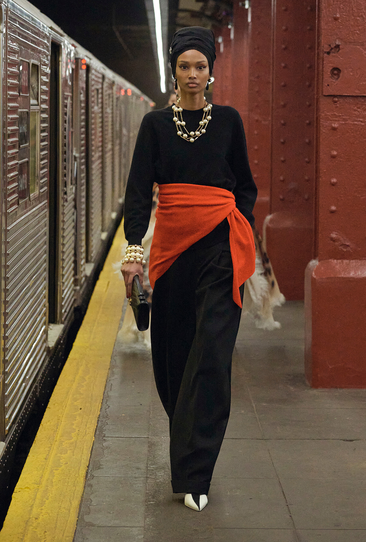 Woman wearing a black outfit with an orange sash walking on a subway platform, headwrap and chunky pearl jewelry.