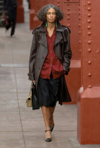Woman walking on a city sidewalk in a long brown leather trench, burgundy blouse, black skirt, green earrings, carrying a beige handbag.