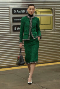 Model in emerald green textured suit with silver trim stands on a subway platform, carrying a two-tone handbag and wearing bold earrings.