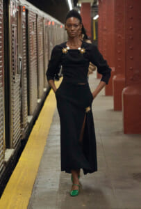 Model in a black midi dress with gold sunburst brooches stands on a subway platform with a yellow edge, hands in pockets.