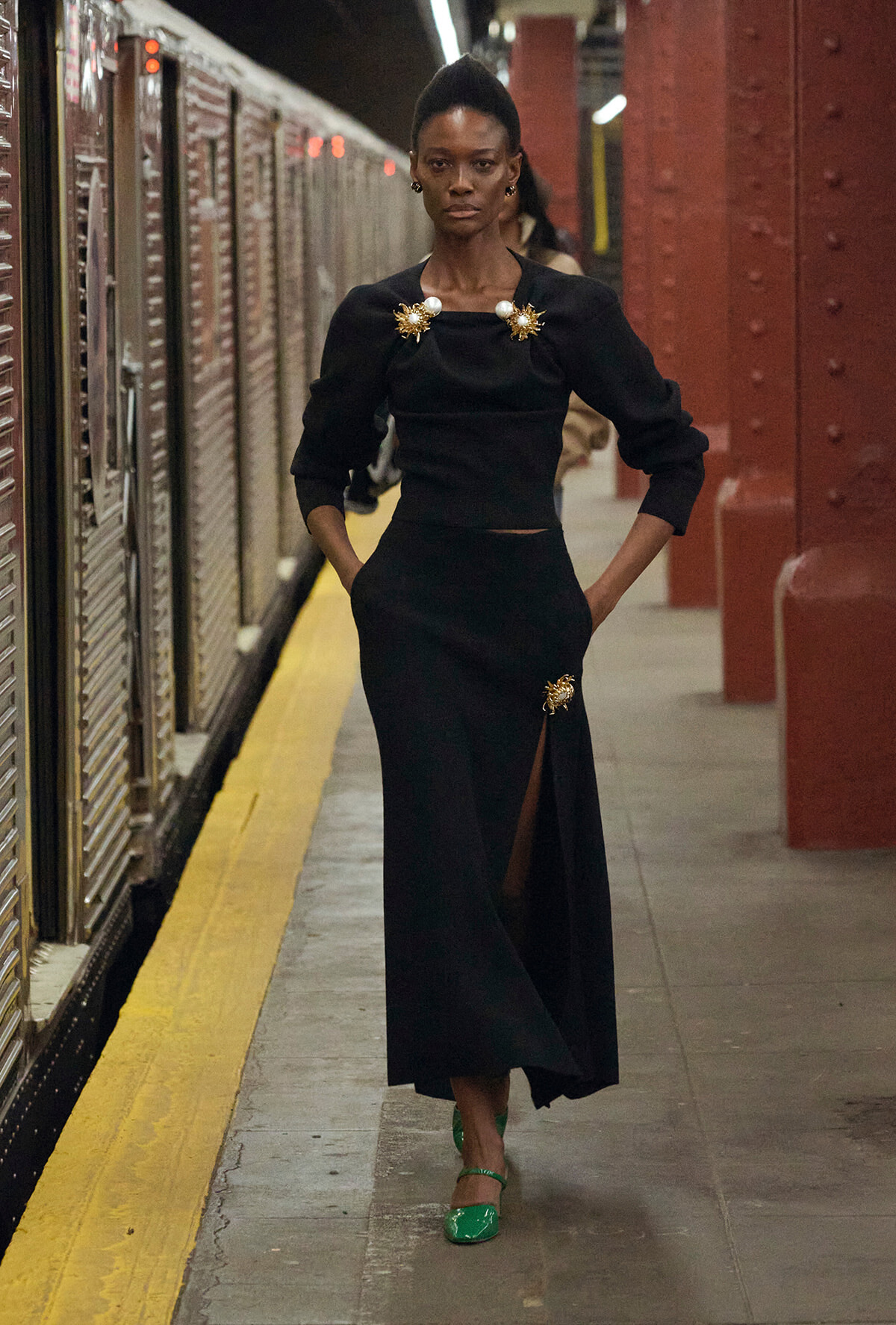 Model in a black midi dress with gold sunburst brooches stands on a subway platform with a yellow edge, hands in pockets.