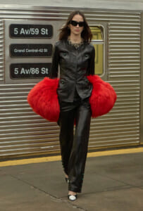 Woman in a black leather suit with a bright red faux fur muff, sunglasses, and statement jewelry on a subway platform.