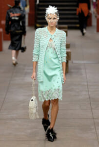 Model walking a runway in a mint lace dress and textured cardigan with a white feathered headpiece and veil, carrying a beaded fringe bag.