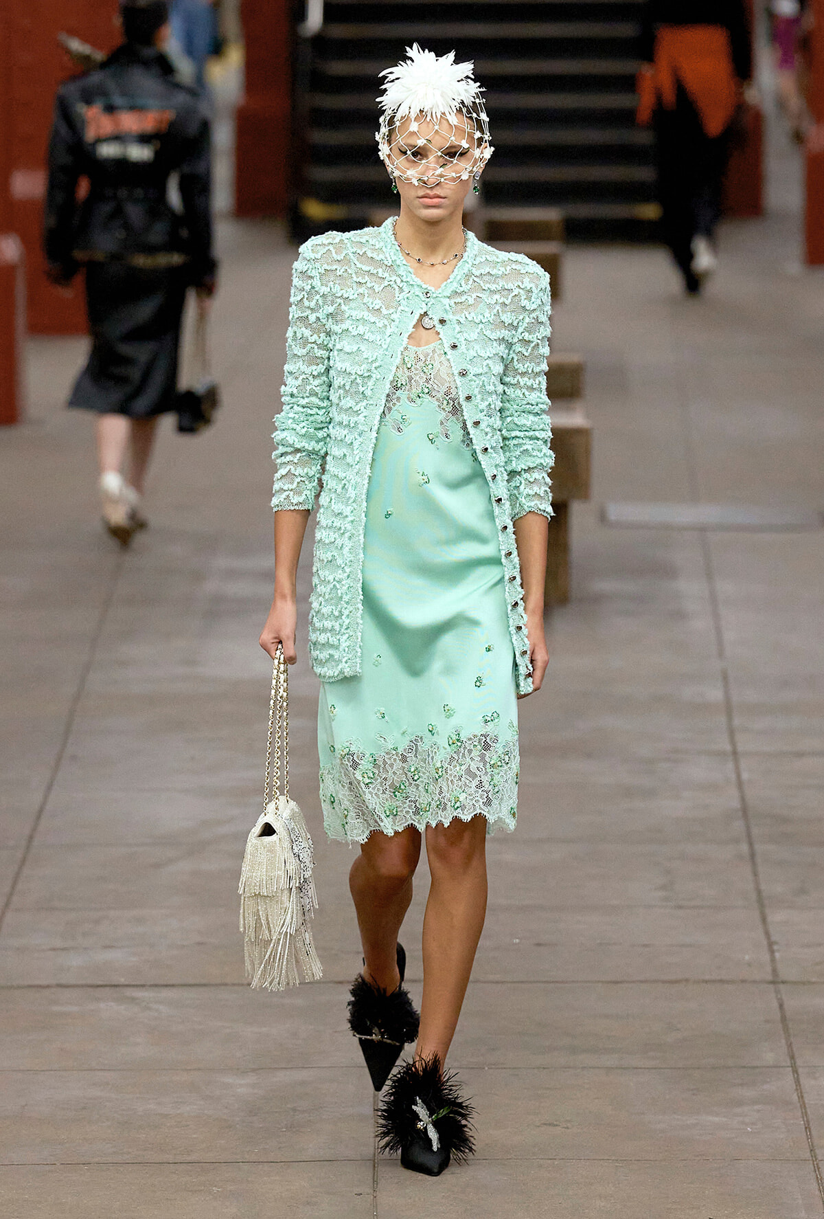 Model walking a runway in a mint lace dress and textured cardigan with a white feathered headpiece and veil, carrying a beaded fringe bag.