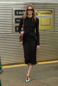 Woman in a black lace midi dress with sunglasses stands on a subway platform, carrying a burgundy handbag and wearing turquoise heels.