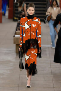 Model on a runway wearing an orange abstract-print dress with black fringe trim, a black turtleneck, white-toe heels, and a black handbag.