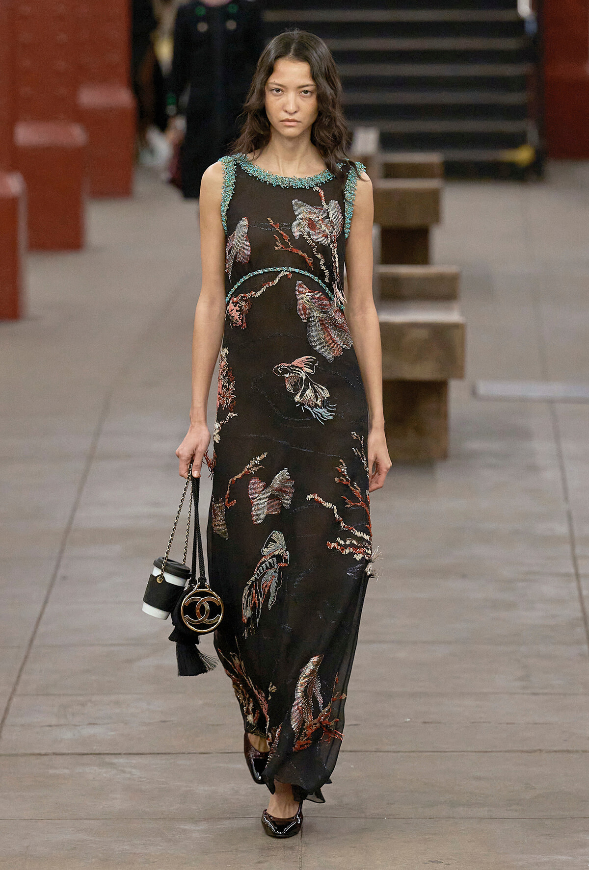 Model walks a runway in a black, floor-length gown with embroidered floral motifs and turquoise neckline embellishments, carrying a small handbag with a chain strap.