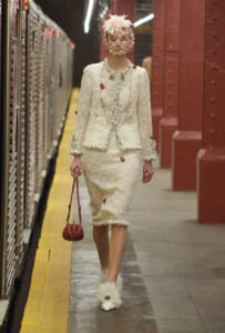Model walking on a subway platform in a cream tweed suit with fringe details, net fascinator, and feathered shoes, carrying a small red handbag.