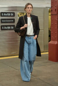 Woman in a black long coat, white top, and wide-leg jeans stands on a subway platform with a brown chain-strap bag.