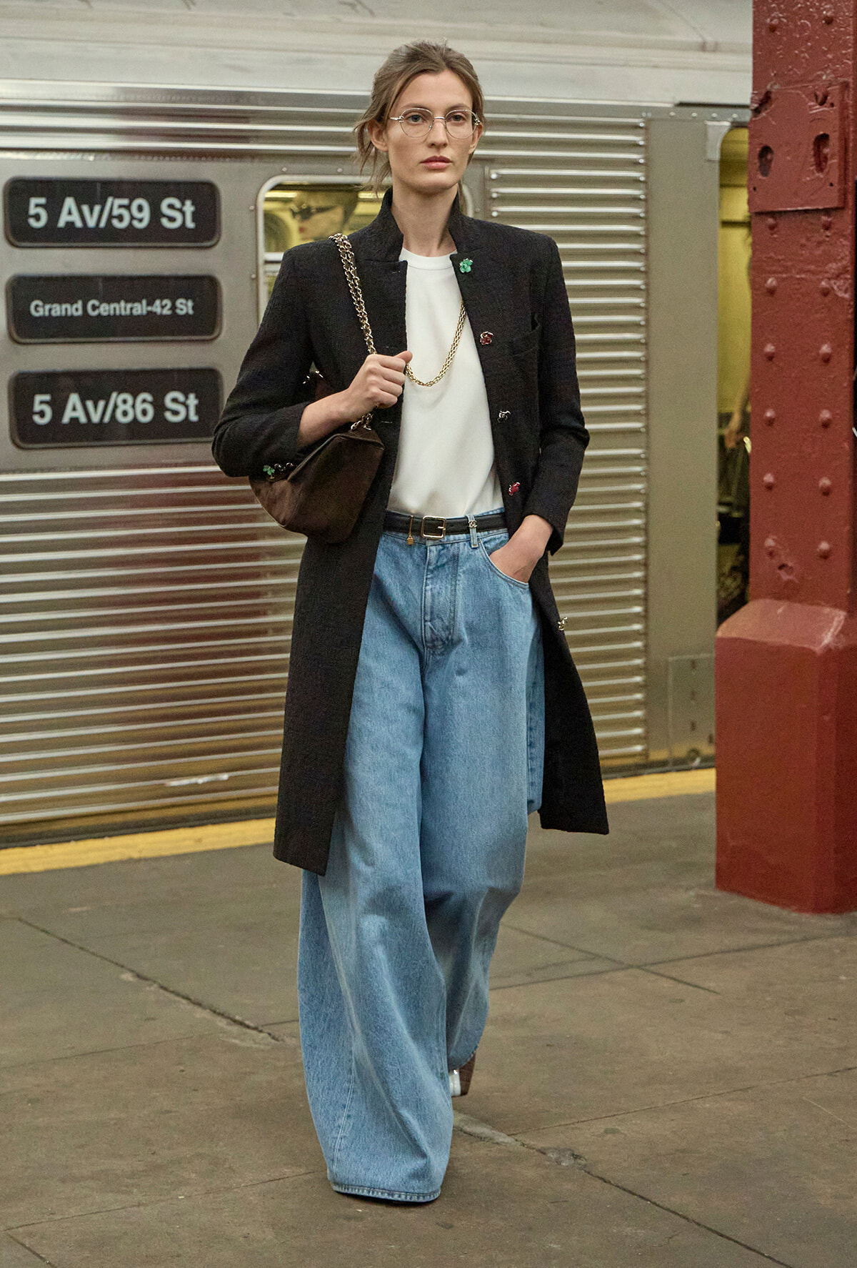 Woman in a black long coat, white top, and wide-leg jeans stands on a subway platform with a brown chain-strap bag.