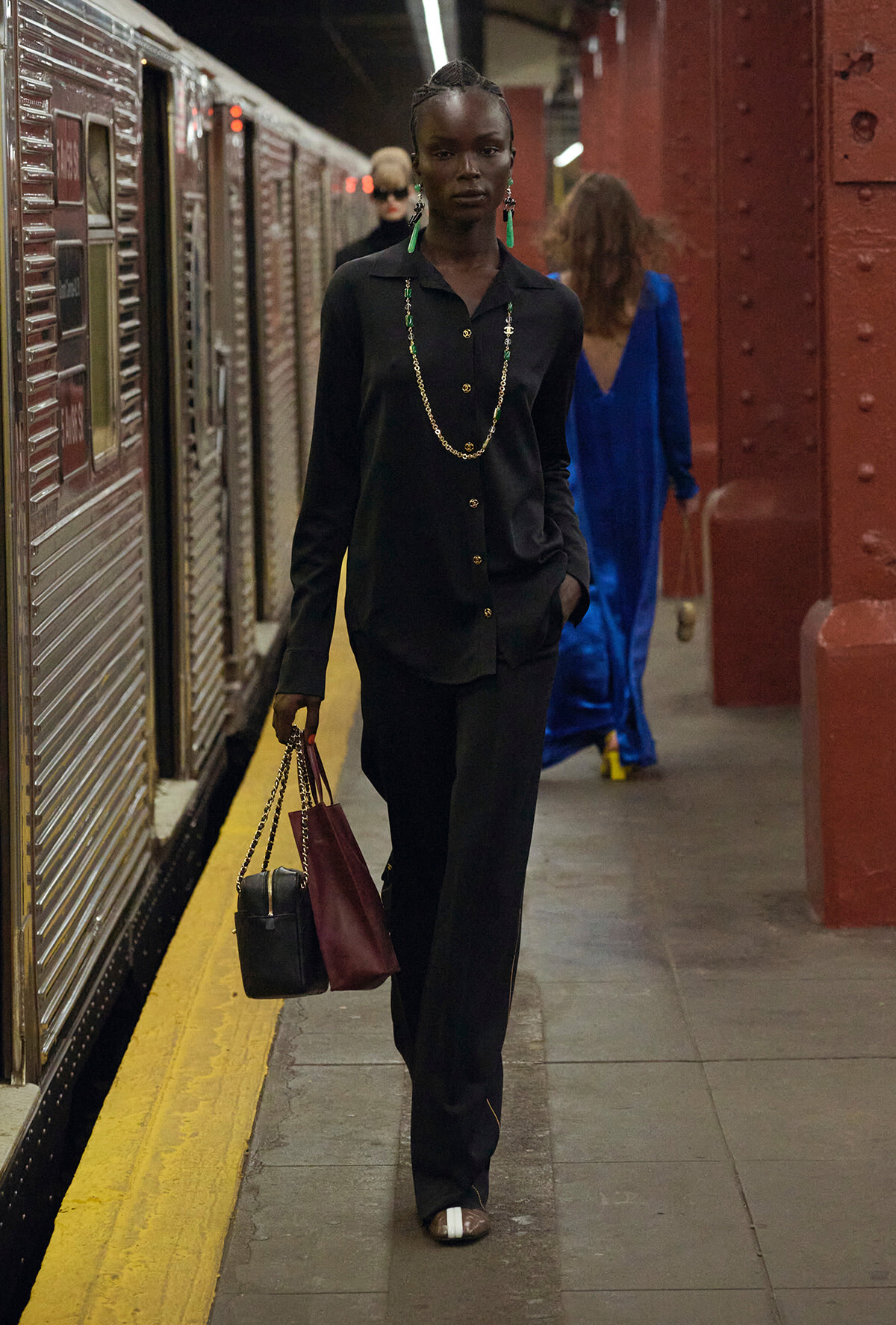 Model in a black pantsuit walking along a subway platform, carrying two handbags and wearing a long gold chain necklace with green earrings.
