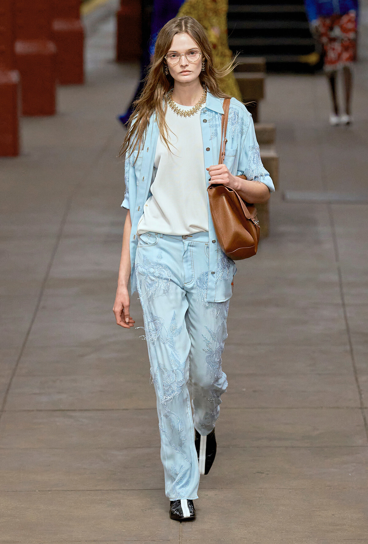 Model walks a runway in a light blue embroidered suit with a white top and brown shoulder bag. She wears glasses and statement jewelry.