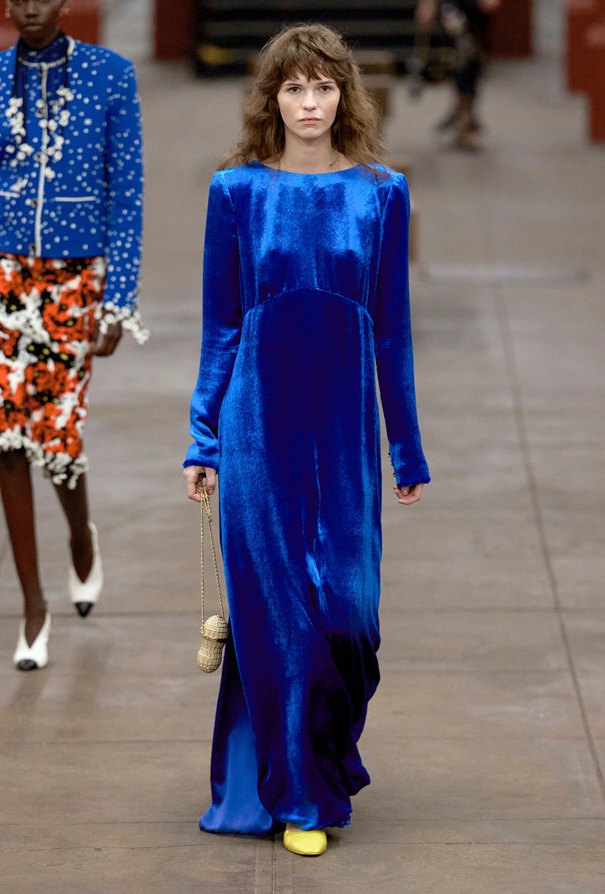 Female model walking a fashion runway in a cobalt blue velvet long-sleeve maxi dress, carrying a small tan woven purse; yellow shoes peek out.