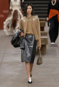 Runway model in a camel embroidered coat over a beige top, paired with a grey crocodile‑texture pencil skirt, white‑and‑black heels, carrying a black coat and a taupe quilted bag.