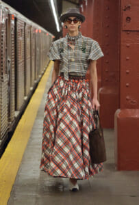 Woman in a gray plaid short-sleeve blouse and long red plaid skirt, wearing a hat, sunglasses, jewelry, and carrying a dark tote on a subway platform.