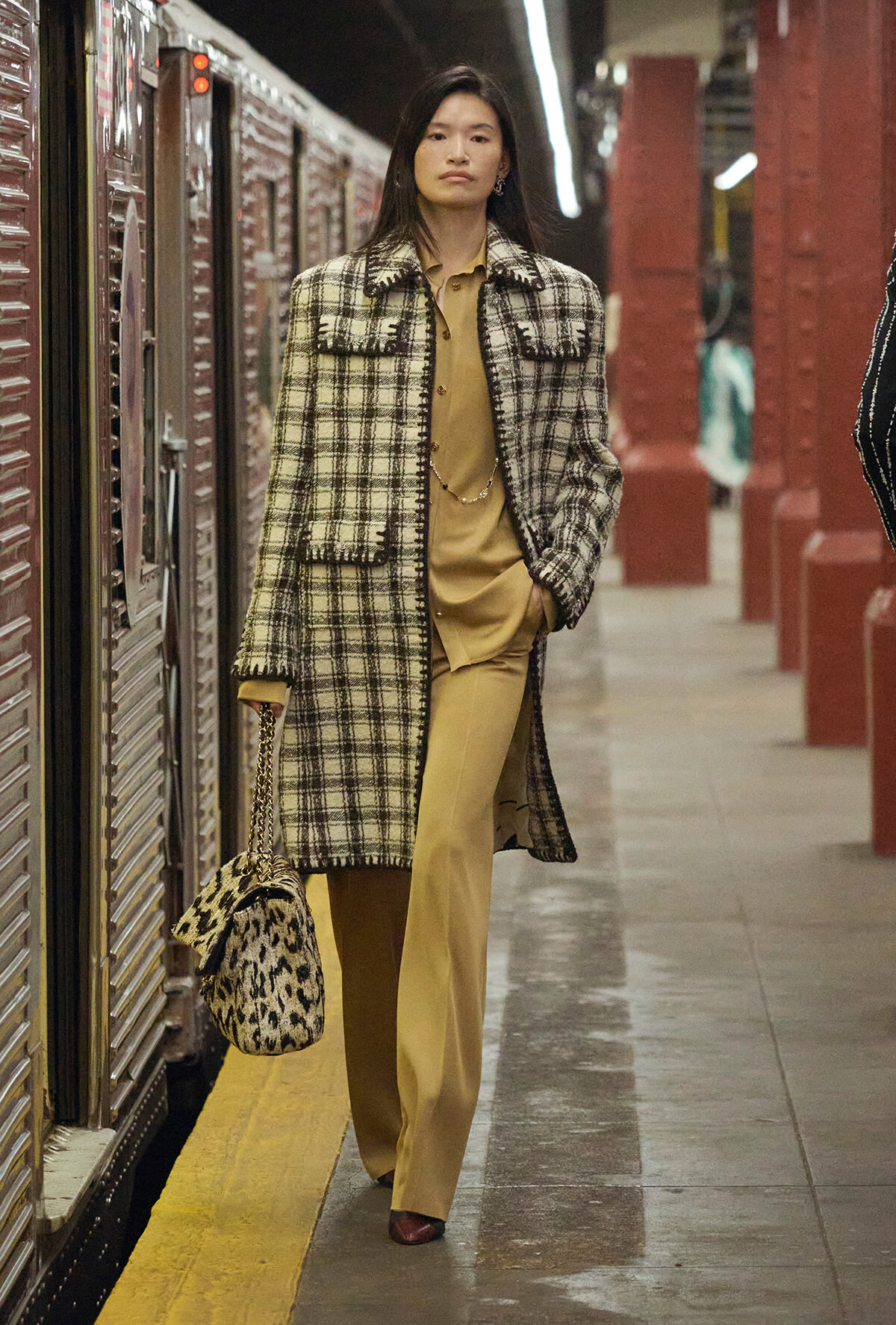 Woman in a beige plaid coat over a tan outfit, carrying a leopard‑print handbag on a subway platform.