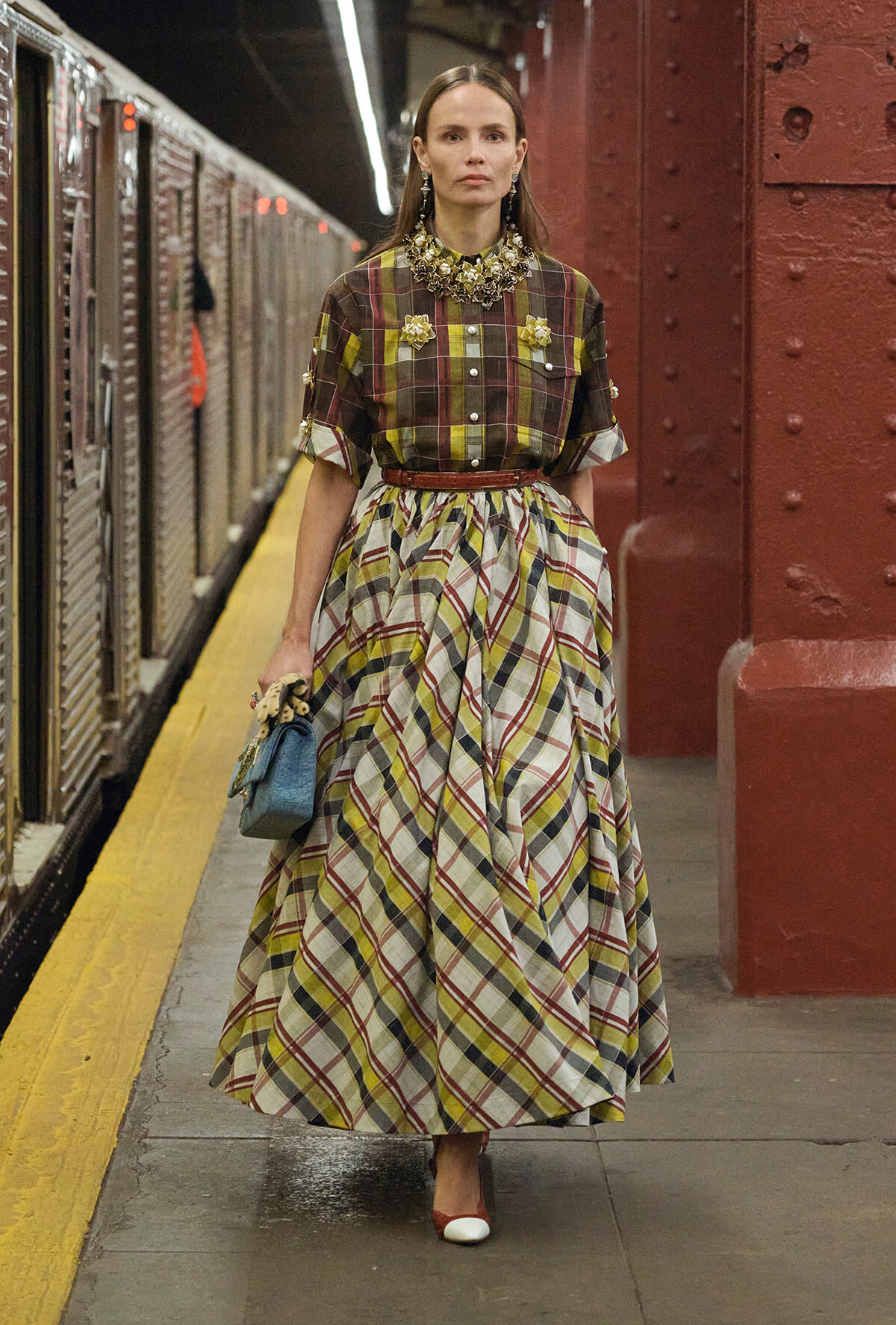 Woman in a brown, yellow, and cream plaid dress with a full skirt stands on a subway platform, holding a small blue handbag and wearing statement jewelry.
