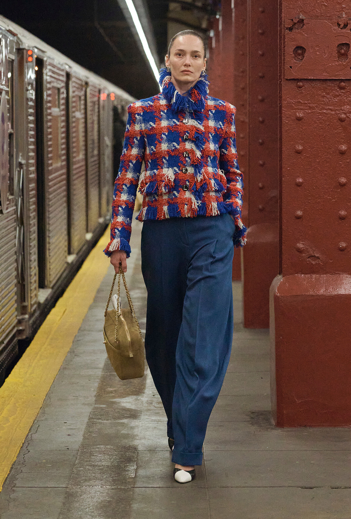 Model walking on a subway platform in a red‑blue fringed blazer, blue wide‑leg pants, and a beige handbag.