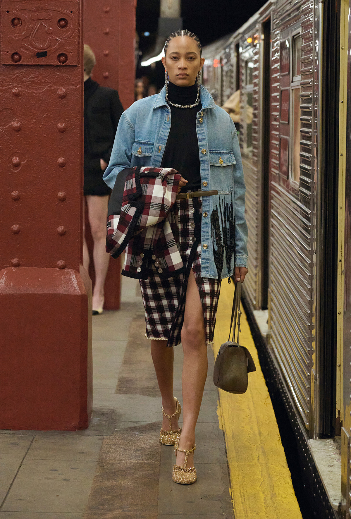 Woman walking on a subway platform in a denim jacket over a black turtleneck, plaid skirt, and glittery gold heels, carrying a plaid coat and a small handbag.