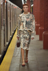 Model in a textured, floral-printed suit with white fringe trim walking on a subway platform, carrying a small brown handbag with a chain strap.