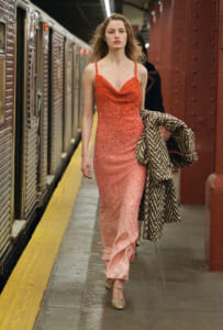 Woman in a sparkling coral-pink evening gown walking along a subway platform, carrying a patterned coat.