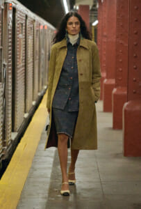Woman with long dark hair and glasses walks along a subway platform, wearing a tan quilted coat over a blue plaid dress and cream shoes.