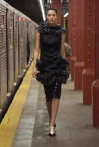 Model walking on a subway platform in a black feather-trim dress with matching heels.