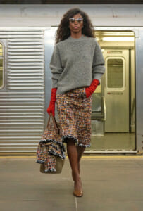 Stylish woman walking on a subway platform in a gray knit sweater, red gloves, a patterned skirt, nude heels, sunglasses, and a large patterned bag. Behind her, an open train door is visible.