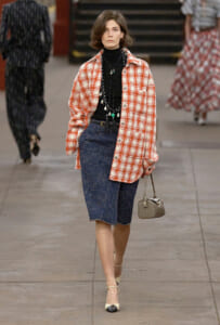 Model walking a fashion runway in a red plaid oversized jacket over a black turtleneck and knee‑length dark denim skirt, accessorized with layered necklaces and a small beige handbag.