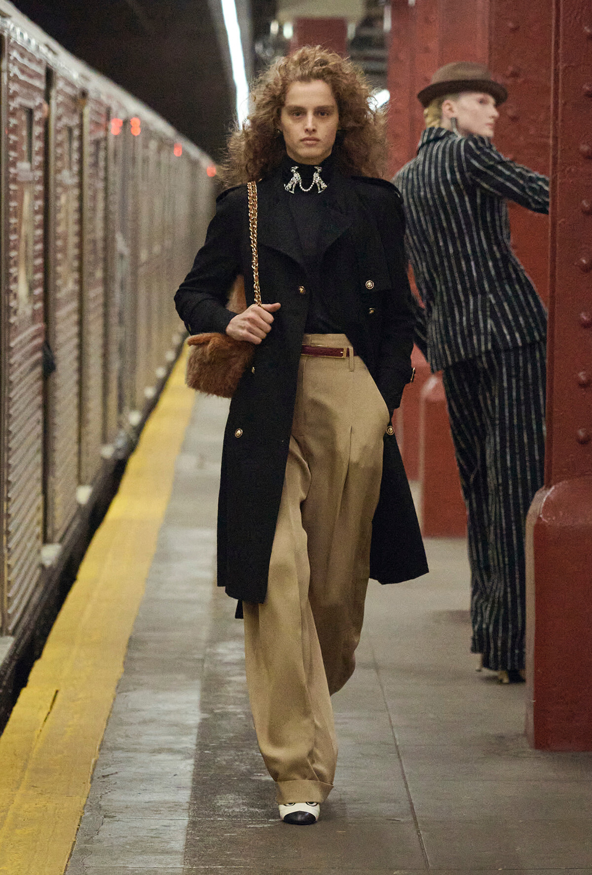 Person walking on a subway platform in a black coat, tan wide-legged trousers, and a chain-strap bag. The scene has a vintage vibe.