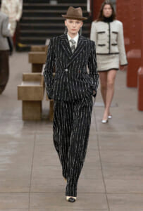 Model on a runway wearing a black pinstripe suit with white vertical stripes, a light shirt and tie, and a brown fedora.