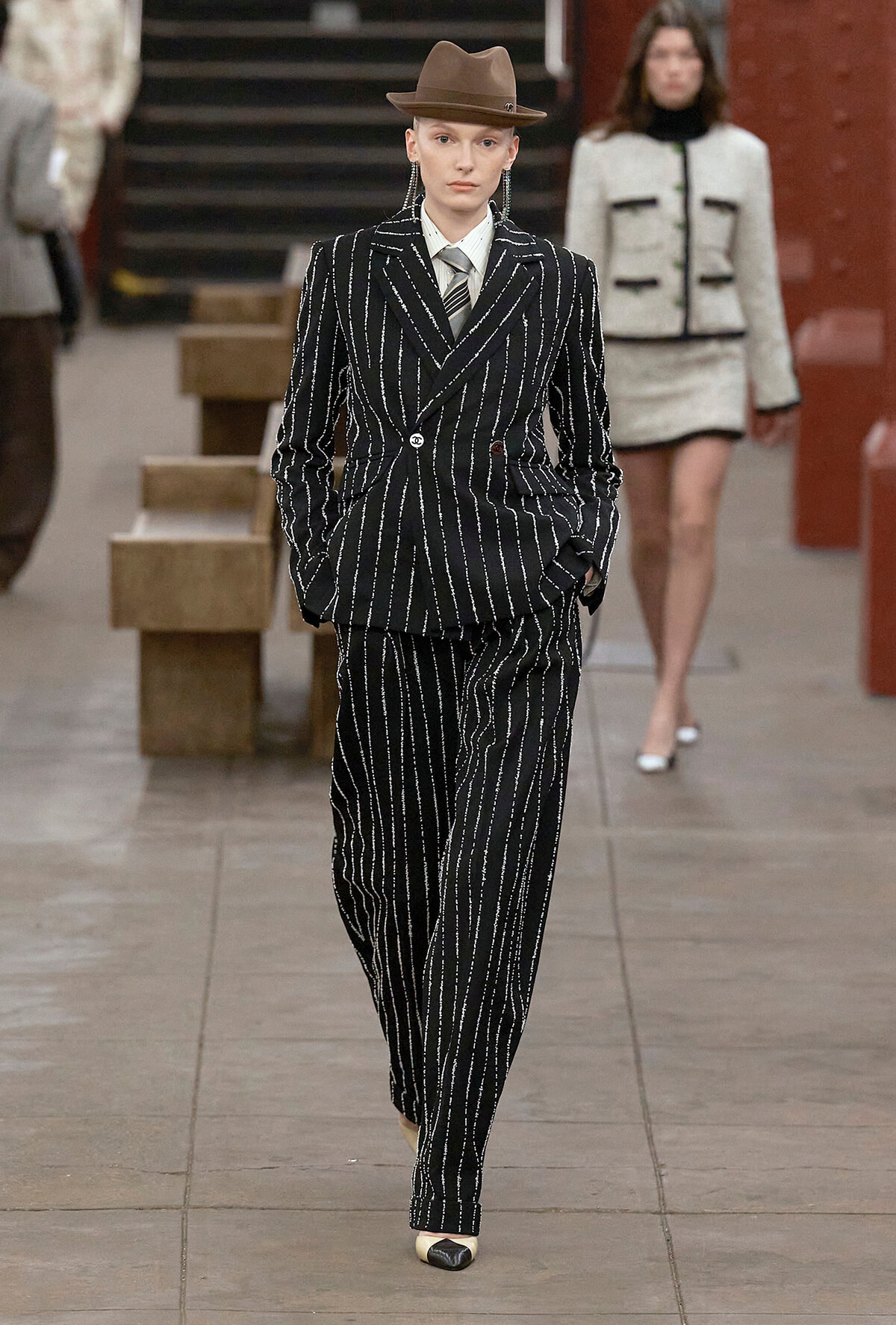 Model on a runway wearing a black pinstripe suit with white vertical stripes, a light shirt and tie, and a brown fedora.