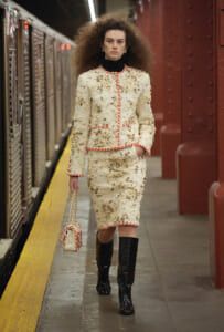 Model walking on a subway platform in a cream embroidered suit with red trim, black turtleneck, and tall black boots, carrying a matching handbag.