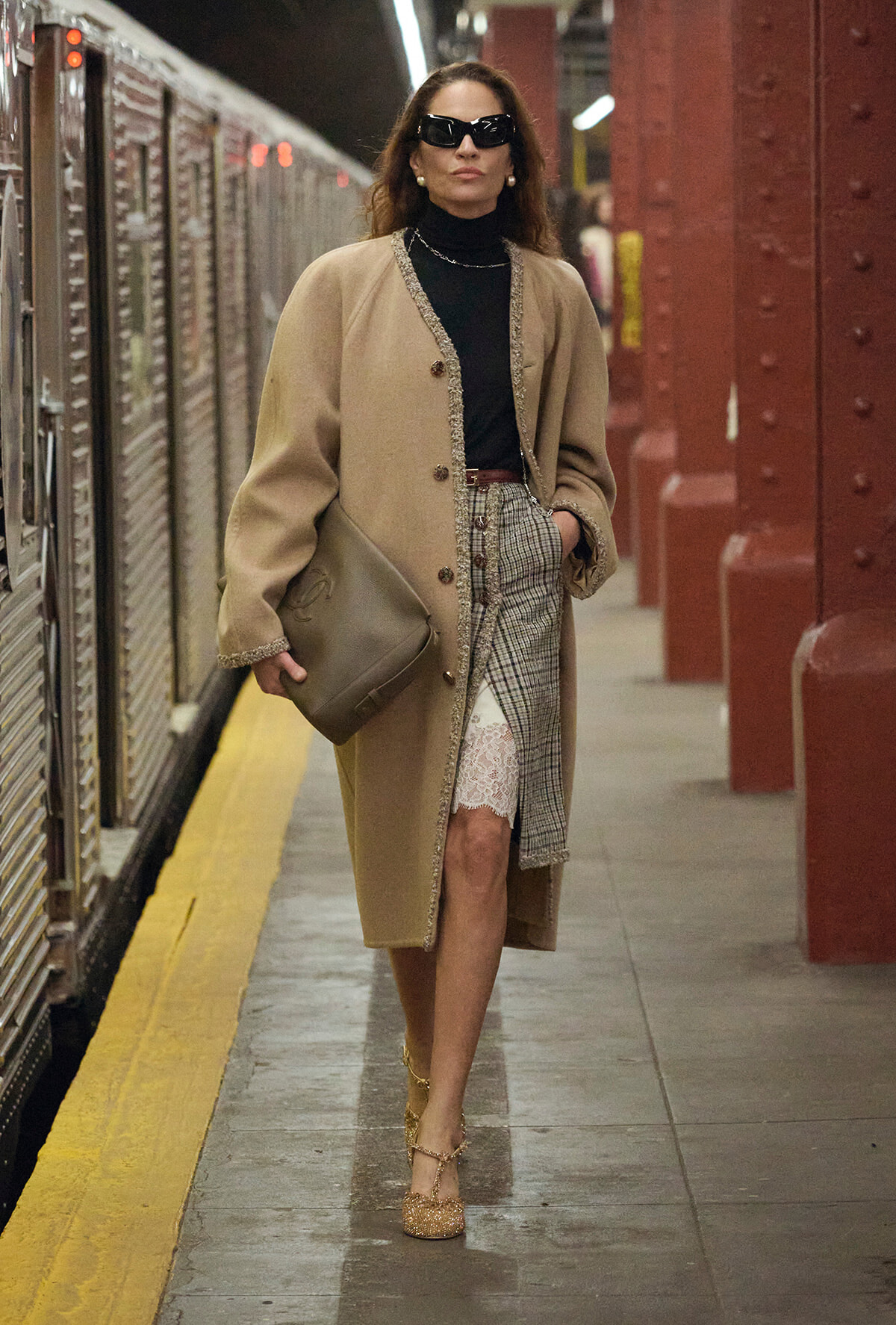 Woman in a beige embellished coat and black turtleneck walking on a subway platform, carrying a taupe clutch and wearing glittery heels.