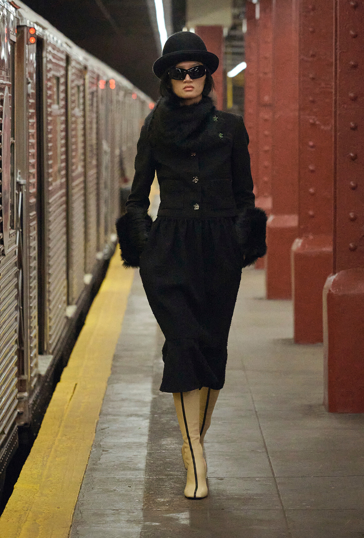 Woman in a black fur-trimmed coat and hat walking along a subway platform, wearing sunglasses.