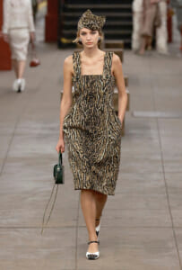 Model walking a runway in a leopard-print sleeveless dress with a matching hat, white heels, and a small green handbag.