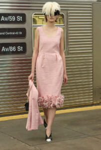Woman in a pink sleeveless dress with a feathered hem walking on a subway platform, holding a matching pink coat and handbag.",