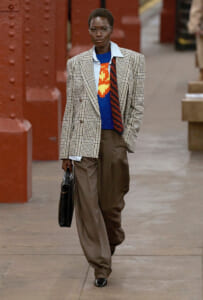 Person walking outdoors in a beige plaid blazer over a blue knit sweater with a yellow-orange pattern, brown wide trousers, glasses, and a black briefcase.