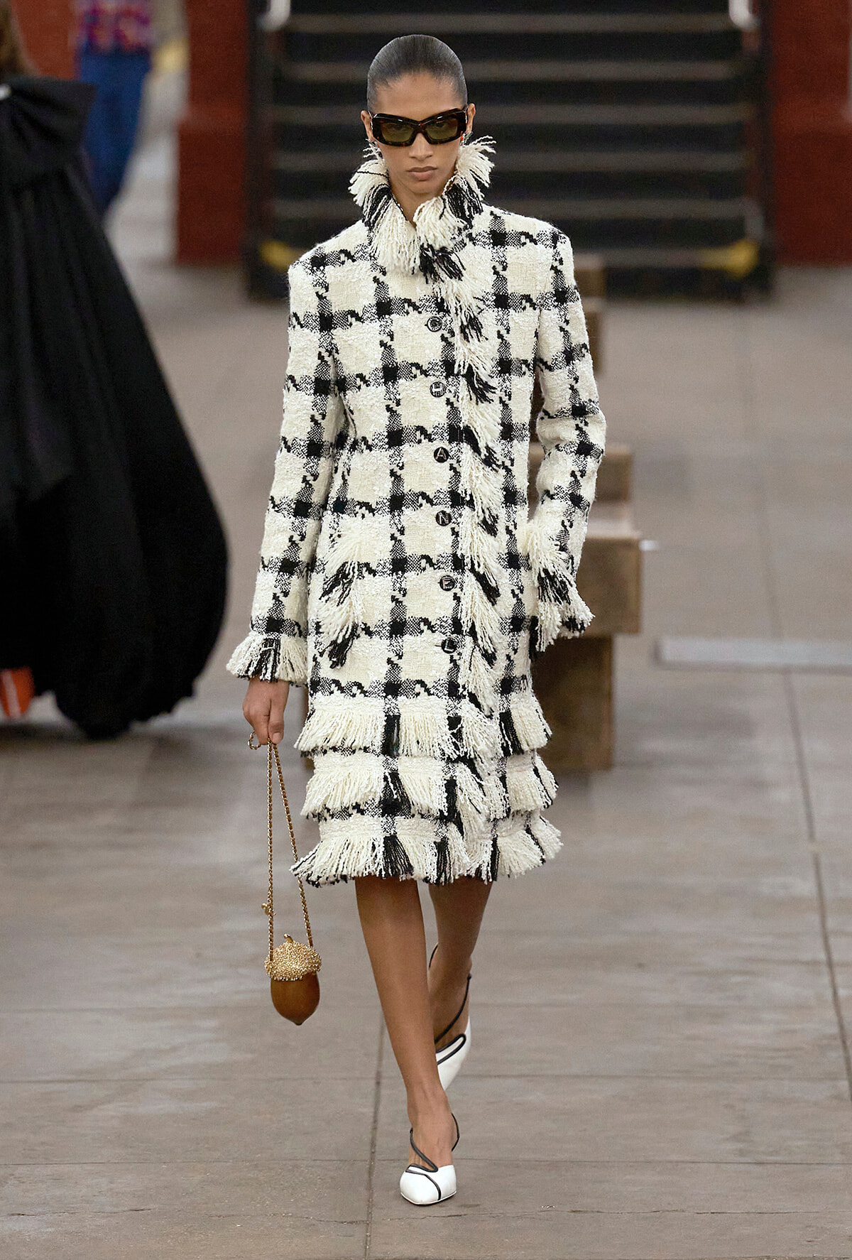 Model wearing a black-and-white checkered tweed coat with fringed edges, black sunglasses, and a small brown beaded handbag, walking on a runway in white heels.