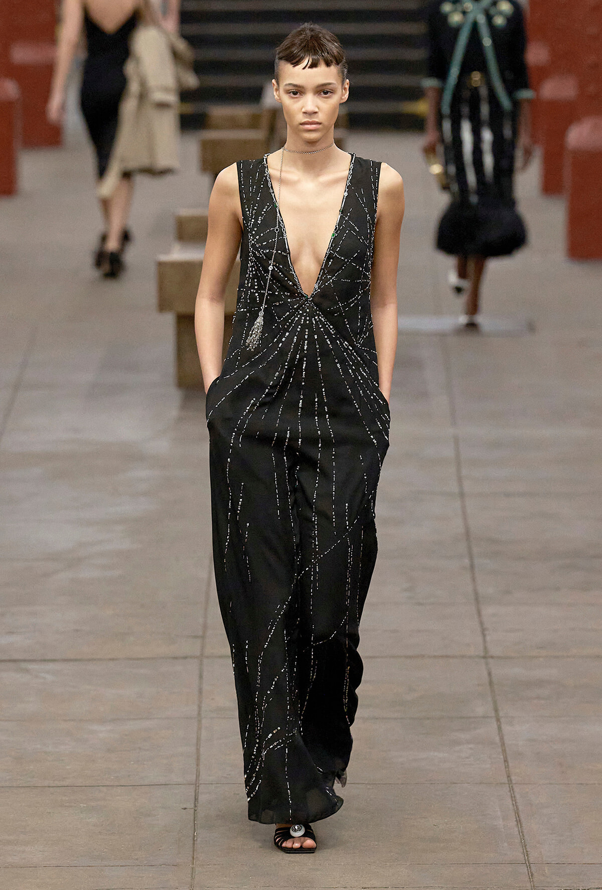 Model on a runway wearing a black plunging-neckline jumpsuit with silver beaded lines and pockets visible in the sides.