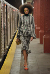Model in a black-and-white tweed suit with green buttons and ruffled peplum walks along a subway platform, carrying a leopard print clutch on her side.