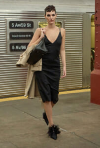 Woman in a black slip dress and trench coat walks on a subway platform, carrying a black handbag and wearing furry-embellished heels.