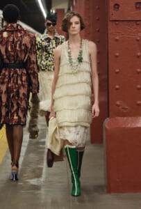 Runway model in a cream tiered fringe dress with green knee-high boots walking beside a red riveted pillar, holding a beige coat.