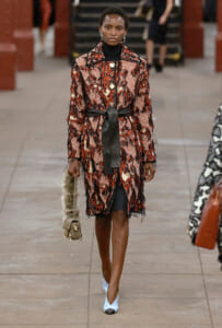 Model on a runway wearing a red-black textured coat with a black sash belt, black turtleneck, and pale blue pointed heels.