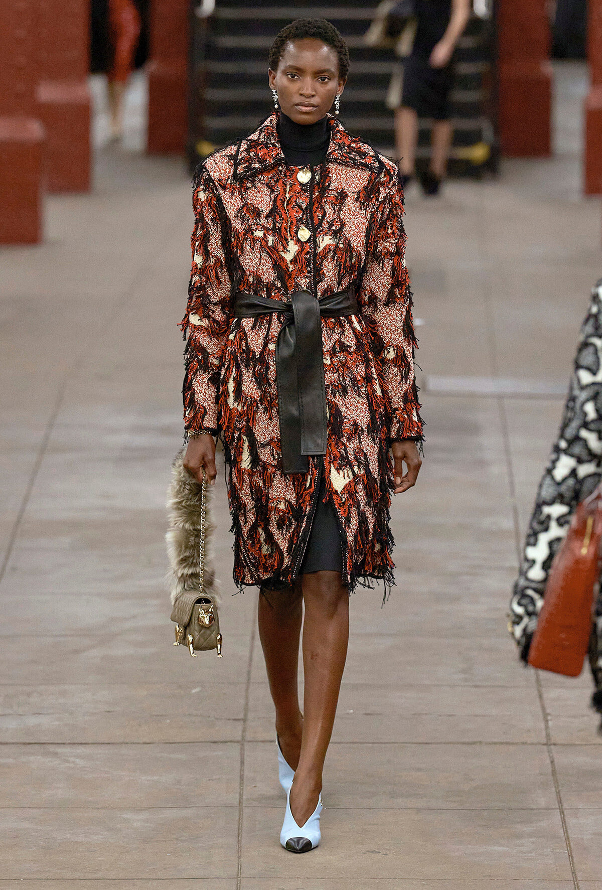 Model on a runway wearing a red-black textured coat with a black sash belt, black turtleneck, and pale blue pointed heels.