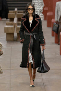 Model wearing a black belted coat with red trim over a lace white dress, carrying a black handbag and wearing sunglasses on a runway session.